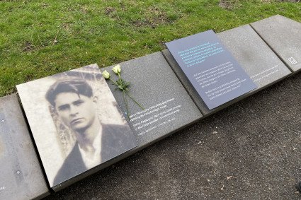 Gedenkstein am Wegesrand mit einem Foto von Spiros Pasaloglou und einem kurzen Text mit seinen Lebensdaten sowie einem Zitat in deutsch und englisch. Auf dem Gedenkstein liegen zwei weiße Rosen.